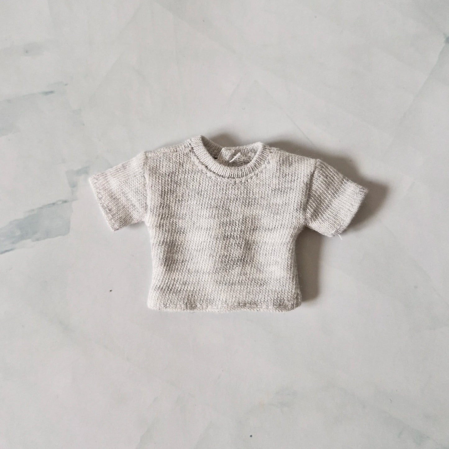 Light grey heather knit short-sleeve crop top for Barbie doll, laid flat on marble background.