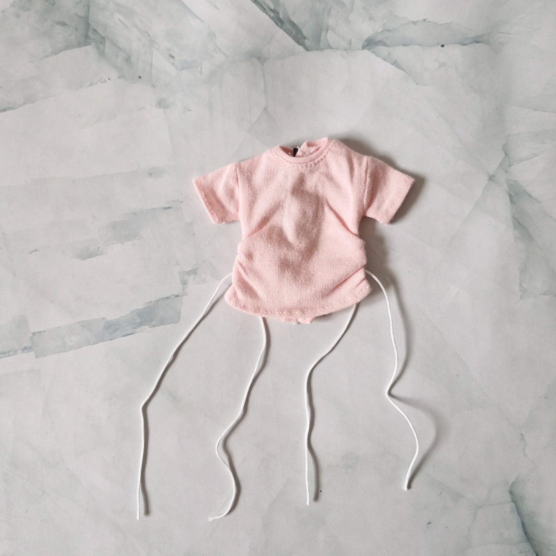Pale pink textured short-sleeve top for Barbie doll with long white drawstrings, laid flat on marble background.