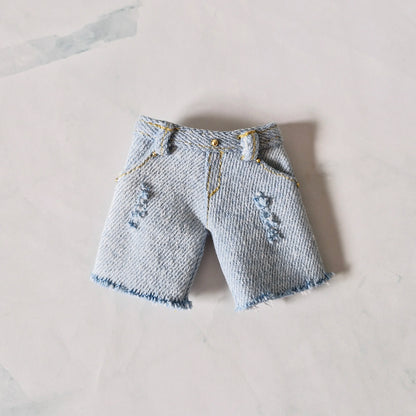 Light wash distressed denim shorts for Barbie doll with frayed hems and gold stud details, laid flat on marble background.