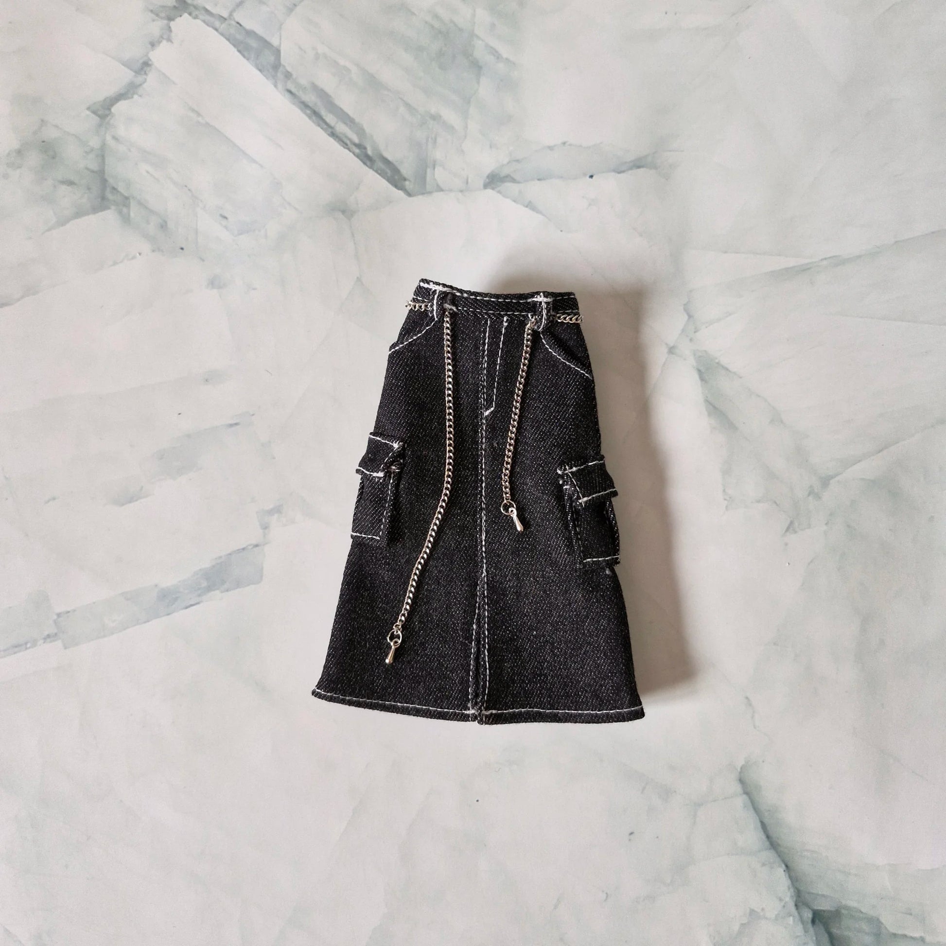 Flat lay of black denim midi cargo skirt with white contrast stitching and silver chain belt detail for fashion dolls on a marble background.